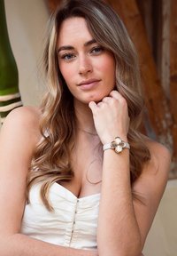 Montre à bracelet blanche avec un accent floral doré. Le bracelet est lisse, et la fleur a une surface douce et brillante. Le modèle pose avec la main sur le menton.