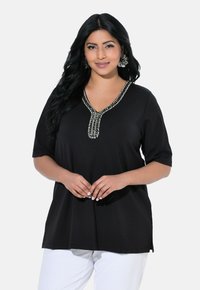 Schwarzes, langgestrecktes Tunika-Oberteil mit V-Ausschnitt und aufwendiger weißer Perlenstickerei am Kragen; kurze Ärmel und weicher, strukturierter Stoff.