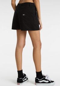 Shorts neri in cotone con una vestibilità rilassata, dotati di una tasca posteriore e un'etichetta del marchio. Indossati con sneaker e calze bianche e nere.