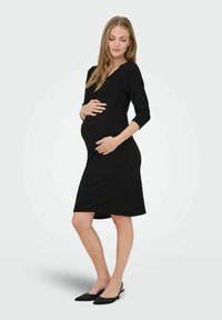 Crna haljina za trudnice s tri četvrtine rukavima, uskim siluetom, mekanim materijalom i dugom do koljena. Nosila se s crnim ravnim cipelama na špic.