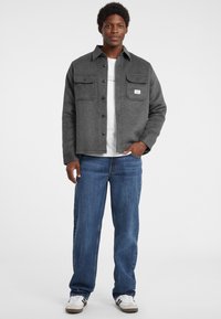 Giacca camicia grigia in misto lana con due tasche sul petto, bottoni neri e una t-shirt bianca a collo rotondo sotto. Indossata con jeans blu e sneakers bianche.