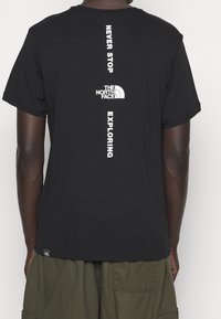 Persona con una camiseta negra de North Face que lleva impresa verticalmente en la parte de atrás el texto "Never Stop Exploring" y el logo, combinada con pantalones cargo oliva.