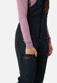 Zwarte skibroek met ritszakken, verstelbare bandjes en het logo "Swedemount". Draagt over een lichtroze lange mouwen top.