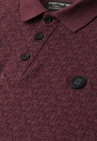 Polo bordeaux avec motif sombre de palmier, trois boutons noirs et un badge logo circulaire noir sur la poitrine.