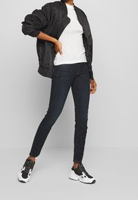 Svart bomberjacka med ribbade ärmar, vit texturerad T-shirt, mörkblå skinny jeans och svarta/vita sneakers, stående mot en neutral bakgrund.