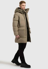Giaccone imbottito in tessuto beige con un grande cappuccio, chiusura con zip e tasche frontali. Pantaloni neri e stivali completano l'outfit.