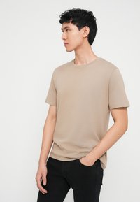 Jeune homme portant un t-shirt beige à manches courtes et un pantalon noir, debout avec une main dans la poche, regardant vers sa gauche sur un fond blanc.