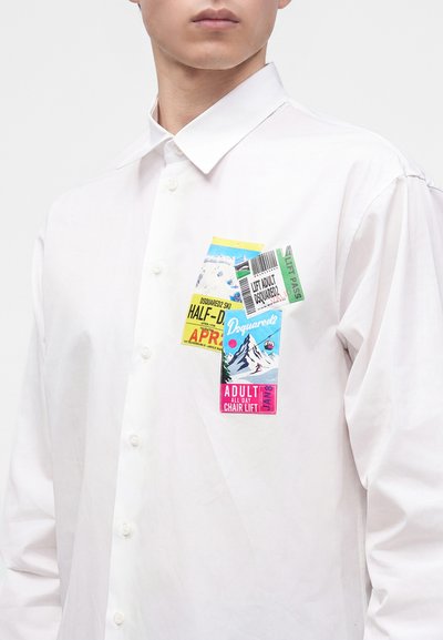 Jeune personne portant une chemise blanche boutonnée avec des motifs colorés de forfait de remontée mécanique et ticket de montagne sur la zone de la poche poitrine.