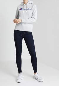 Sweatshirt à capuche gris avec des cordons blancs, arborant un logo "Champion" brodé en bleu et rouge. Porté avec un jean foncé et des baskets blanches.