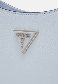 Lyseblå håndtaske med tekstureret materiale, prydet med en trekantet gylden og hvid logo med teksten "GUESS USA" og et spørgsmålstegn som detalje.