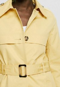Trench-coat jaune pâle avec un col pointu, une fermeture centrale à boutons et une ceinture assortie avec une boucle carrée. Détail de tissu texturé visible.