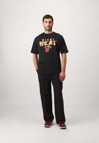 Camiseta negra con un colorido gráfico de "MIAMI HEAT", combinada con pantalones negros y zapatillas con acentos rojos. Look simple y casual.