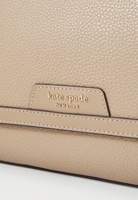 Geantă de mână din piele bej cu un finisaj texturat, având un logo auriu "kate spade NEW YORK" și cusături contrastante pe bretele.