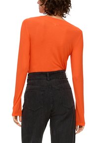 Langarm, leuchtend orange Oberteil kombiniert mit hochgeschnittenen, dunkelgrauen Jeans, die über Taschen und ein Hüftbundetikett verfügen.