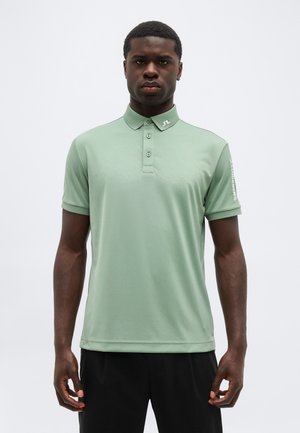 Mann trägt hellgrünes Poloshirt mit kurzen Ärmeln, geknöpftem Kragen und Logo am Kragen sowie am linken Ärmel, steht vor einfachem Hintergrund.