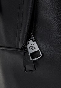 Détail de la fermeture éclair d'un sac en cuir noir avec deux tirettes métalliques noires. Chaque tirette est ornée d'un logo "cK" embossé. Texture subtilement visible sur le cuir.