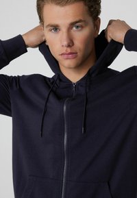 Námořnická mikina na zip s kapucí na šňůrku, klokankovými kapsami a texturovanou tkaninou. Model si stáhne kapuci dozadu, čímž odhalí hladký materiál a střih.