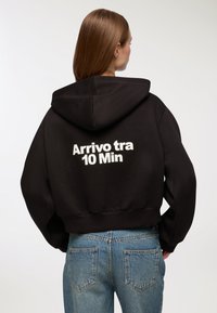 Felpa nera a vita corta con una grande scritta bianca sulla schiena che recita "Arrivo tra 10 Min." Presenta un cappuccio con cordini e una vestibilità comoda.