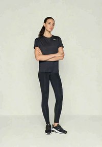 Ženska stoji s prekriženimi rokami, oblečena v črno Nike trenirko, pajkice in črne športne copate, pred enobarvno svetlo ozadjem.