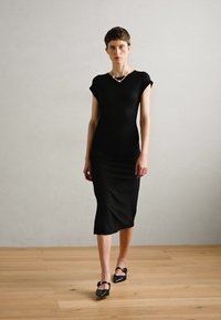Vestido negro ajustado de manga corta, con dobladillo a la altura de la rodilla, de textura suave. Combinado con zapatos planos negros con una correa decorativa.
