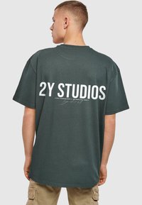 Tmavě zelené oversized tričko má na zadní straně velký bílý nápis "2Y STUDIOS" a žebrovanou texturu. Krátké rukávy a kulatý výstřih.