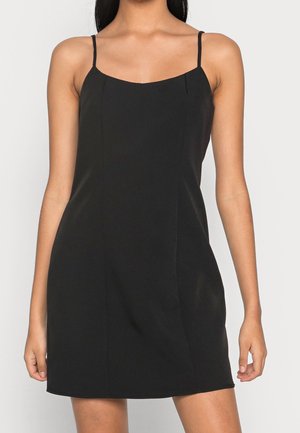 Donna che indossa un mini abito nero senza maniche con sottili spalline su uno sfondo chiaro e uniforme.