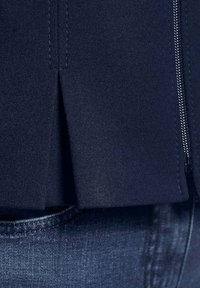 Marineblaues Kleidungsstück aus strukturiertem Stoff, mit vertikalem Schlitzdetail und Reißverschluss, kombiniert mit dunklen Jeans.