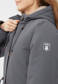Šedá nepromokavá bunda se strukturálním povrchem, kapucí podšitou kožešinou, šňůrkou na stažení a logem na levém rukávu.