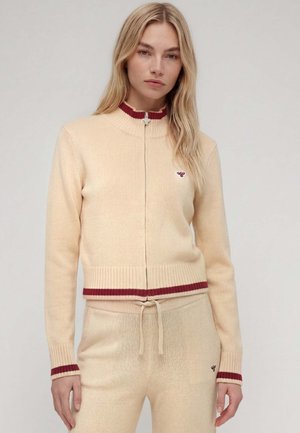 Jeune femme portant un pull en maille beige à fermeture éclair et un pantalon assorti avec des bordures rouges aux poignets, au col et à la taille sur fond uni.
