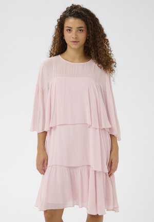 Giovane donna con capelli ricci che indossa un vestito rosa chiaro a strati con maniche trasparenti, in piedi davanti a uno sfondo bianco semplice.