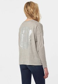 Grijze langemouwen sweater met een glanzend zilveren grafisch ontwerp op de achterkant. Zachte textuur en een relaxte pasvorm, getoond met donkere jeans.