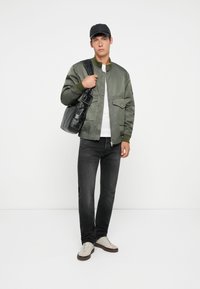 Giacca bomber verde oliva con tasche frontali, una maglietta bianca sotto, jeans scuri e sneakers beige. Una borsa nera e un cappellino completano il look.
