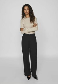 Top beige à manches longues avec un design ajusté, associé à un pantalon noir à jambes larges taille haute et des escarpins noirs à bout pointu.