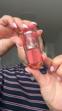 Gjennomsiktig rosa kosmetikkflaske med et gullbånd, som inneholder en ferskenfarget væske. Brukerens hånd delvis synlig, holder flasken.