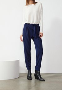 Mörkblå skräddarsydda byxor med ett matchande bälte i samma tyg, kombinerat med en vit långärmad topp, kompletterat med svarta kilklackar vid ankeln.