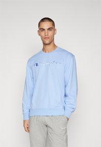 Hellblaues Sweatshirt mit langen Ärmeln, das ein großes gesticktes "Champion"-Logo auf der Brust trägt. Rippbündchen und -saum.