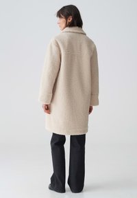 Beige Kunstpelzjacke mit einem breiten Kragen, langen Ärmeln und einem mittel-Längen-Schnitt, getragen mit dunklen Jeans. Weiche Textur und minimalistisches Design.