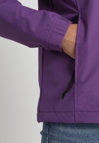 Hånd i sidelommen på lilla jakke med elastisk mansjet og sort snor, båret over blå denimjeans.