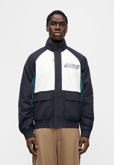 Άνδρας που φοράει μπουφάν Billionaire Boys Club σε σκούρο μπλε, άσπρο και μπλε-πράσινο με μπροστινές τσέπες και μπεζ παντελόνι, στέκεται μπροστά σε απλό φόντο.