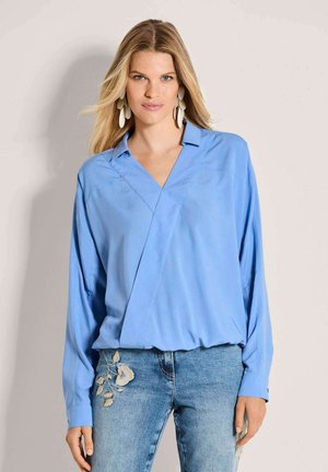 Blonde Frau trägt übergroße hellblaue V-Ausschnitt-Bluse und blaue Jeans mit Blumenstickerei, steht vor schlichtem Hintergrund.