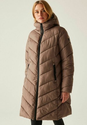 Mujer con un abrigo largo acolchado de color topo con capucha y bolsillos con cremallera negros, de pie frente a un fondo liso.