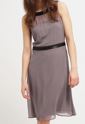 Vestido gris sin mangas hasta la rodilla con ribete de lentejuelas negras en el escote y la cintura, llevado por una mujer con el pelo suelto.