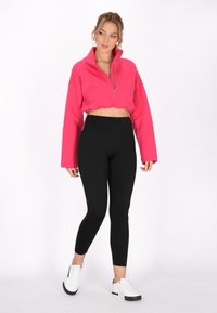 Felpa rosa brillante, tagliata, con colletto alto e zip frontale, abbinata a leggings neri e sneakers bianchi. Design semplice e aderente.