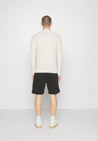 Camisa ligera de manga larga en color crema, combinada con pantalones cortos negros, que presenta un corte relajado y una textura suave. Calzado informal completa el atuendo.