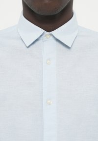 Ανοιχτό μπλε, πουκάμισο με κουμπιά από λινό ύφασμα. Διαθέτει κλασικό γιακά και διαφανή πλαστικά κουμπιά. Μαλακή υφή με διακριτικό, λείο φινίρισμα.