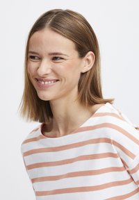Csíkos, hosszú ujjú póló fehér és barack színben, széles nyakkivágással és puha textúrával. A modellt mosolyogva láthatjuk, fülcimpáját is megmutatva.