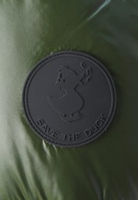 Un tissu vert et brillant présente un badge rond en caoutchouc noir avec une illustration de canard et le texte "SAUVEZ LE CANARD" en bas.