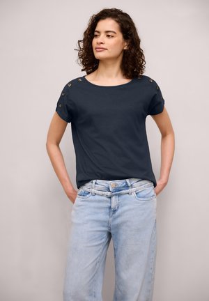 Donna con capelli ricci che indossa una maglietta a maniche corte blu scuro con bottoni sulle spalle e jeans azzurri, con le mani nelle tasche.