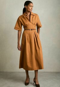 Bruine katoenen midi-jurk met korte mouwen, ceintuurelement en zijzakken. De stof heeft een zachte textuur en een gestructureerd ontwerp.