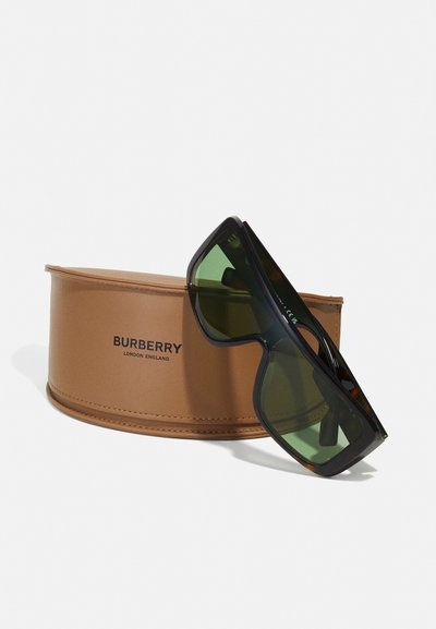 Occhiali da sole Burberry con montatura nera spessa e lenti verde scuro, posizionati su un astuccio circolare beige con il logo Burberry.